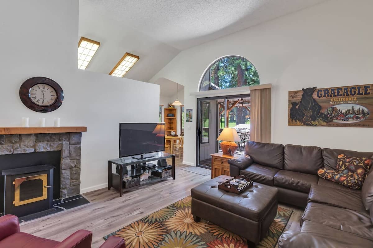 Living room beneath an arched window with a stone-surround wood stove, leather sectional and a slider onto the deck at 121 Cottonwood, Plumas Pines — 3-bedroom Plumas Pines vacation rental in Graeagle, CA